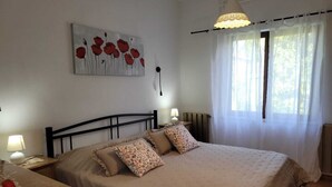 A bedroom with a bed, two bedside tables, a lamp, a radiator, a window with curtains, and a painting on the wall.