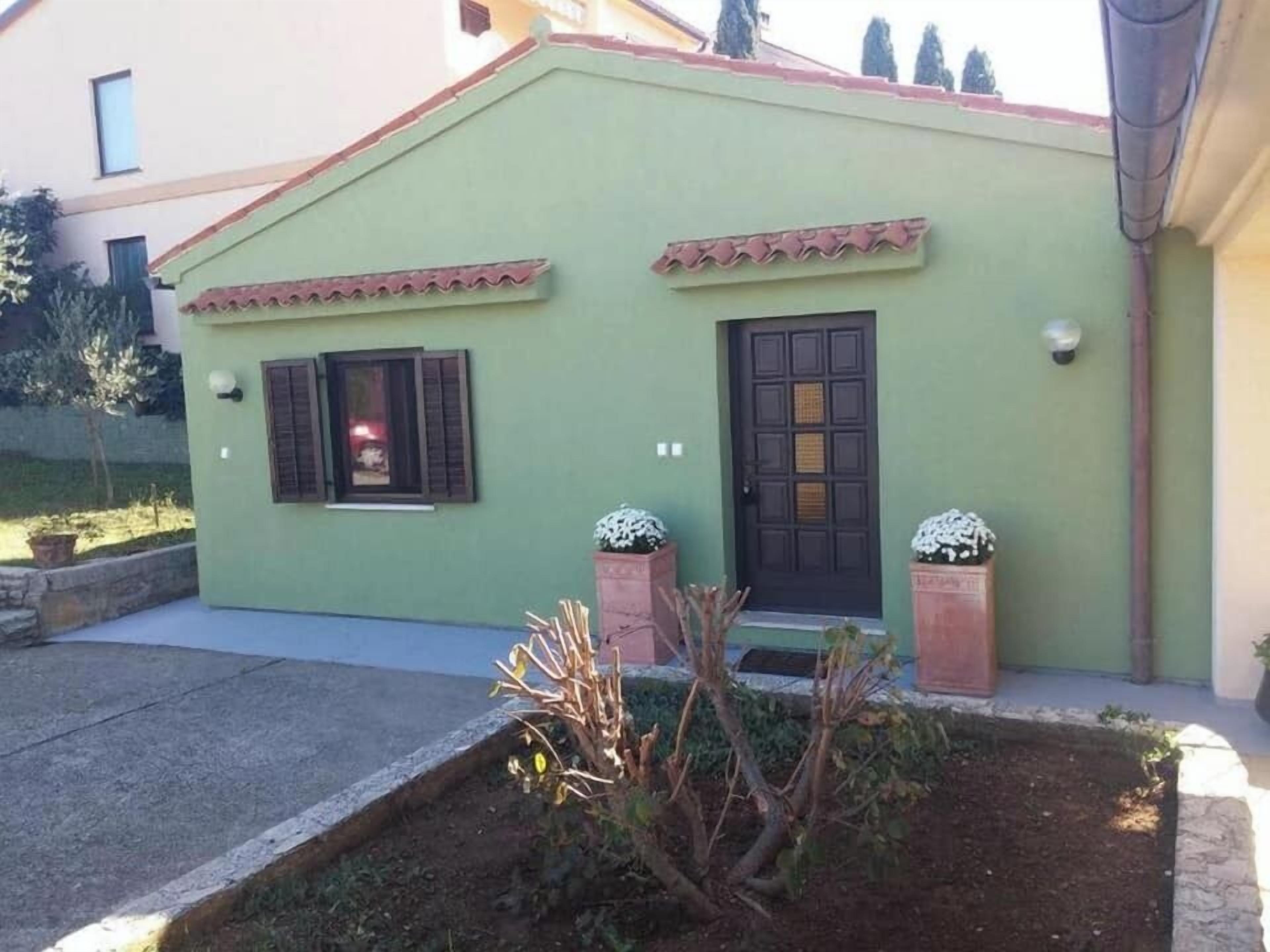 A small, single-story building with a green facade, a black door, and two windows with shutters. There are two potted plants in front of the door.