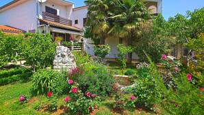 A two-story house with a garden in front, featuring a stone wall, flowering plants, and a palm tree.