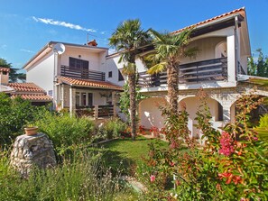 Traditional Mediterranean-style building with a flat roof and balcony, surrounded by lush greenery and a stone feature.