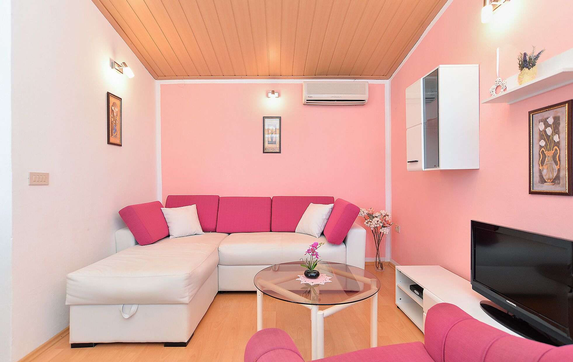 Una sala de estar con pared rosa, sofá blanco, una mesita de centro de vidrio, un televisor de pantalla plana y un armario blanco.