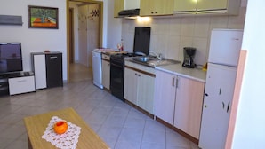 Uma cozinha compacta com fogão, pia e geladeira. Há uma mesa pequena com uma laranja. Uma televisão está sobre um armário à esquerda.