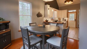 A dining area with a round table and six chairs, a kitchen in the background, and a large window providing natural light.