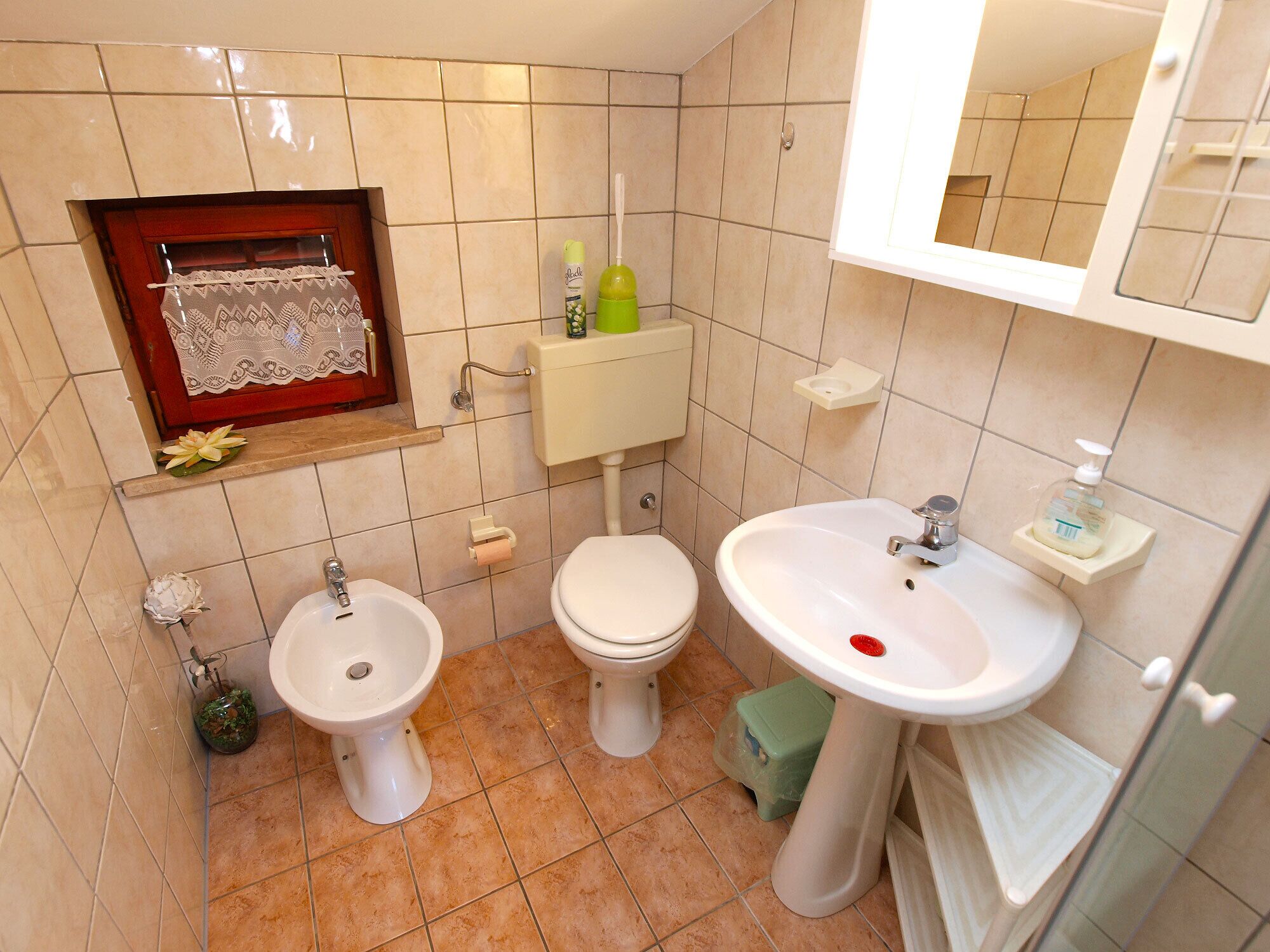 A small bathroom with a toilet, bidet, and sink. There is a mirror above the sink and a shelf with a lace item.