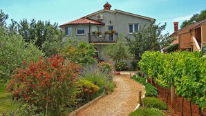 A two-story house with a red-tiled roof, surrounded by a garden with various plants and a gravel path leading to the entrance.