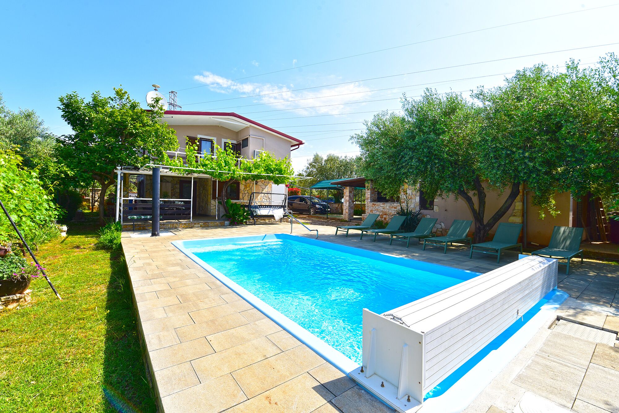 A swimming pool with a white deck, surrounded by a garden with trees and a patio area with lounge chairs.