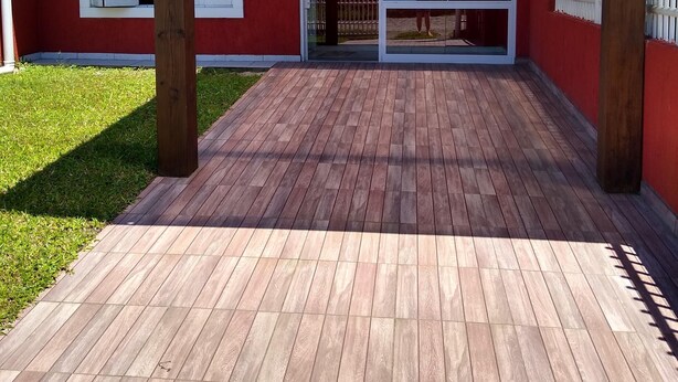 A covered patio area with a wooden deck, a glass door entrance, and a white fence. - Casa Jardim Beira Mar (Capão da Canoa)