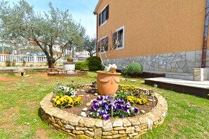 Un giardino con un cordolo in pietra, un vaso centrale con fiori e un edificio con facciata e finestre in pietra.