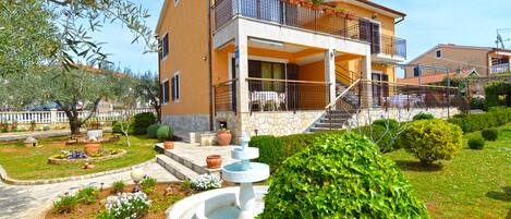 A two-story house with a balcony, a stone staircase, and a garden with a fountain.