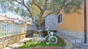 Ein weißes Fahrrad steht unter einem Baum, im Hintergrund eine Steinmauer und ein Metallzaun.