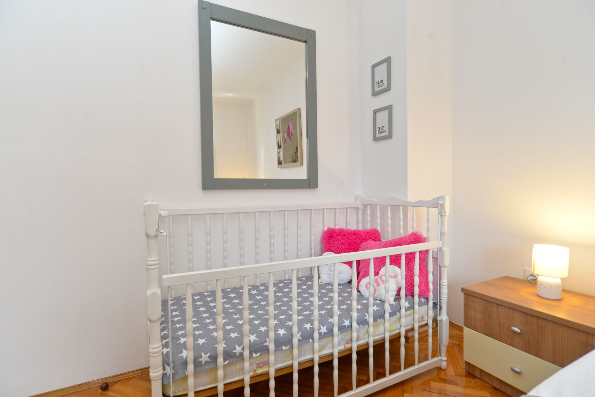 Una cuna para bebé con estructura blanca y colchón gris con estampado de estrellas, un mesita de noche de madera con una lámpara, y un espejo montado en la pared.