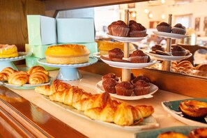 Diversa variedad de pasteles expuestos en bases escalonadas, incluyendo cruasanes, muffins y un pastel de queso.