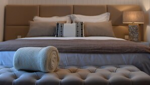 Una cama bien tendida con un cabecero marrón, una lámpara de noche y una toalla enrollada sobre un otoman tapizado.