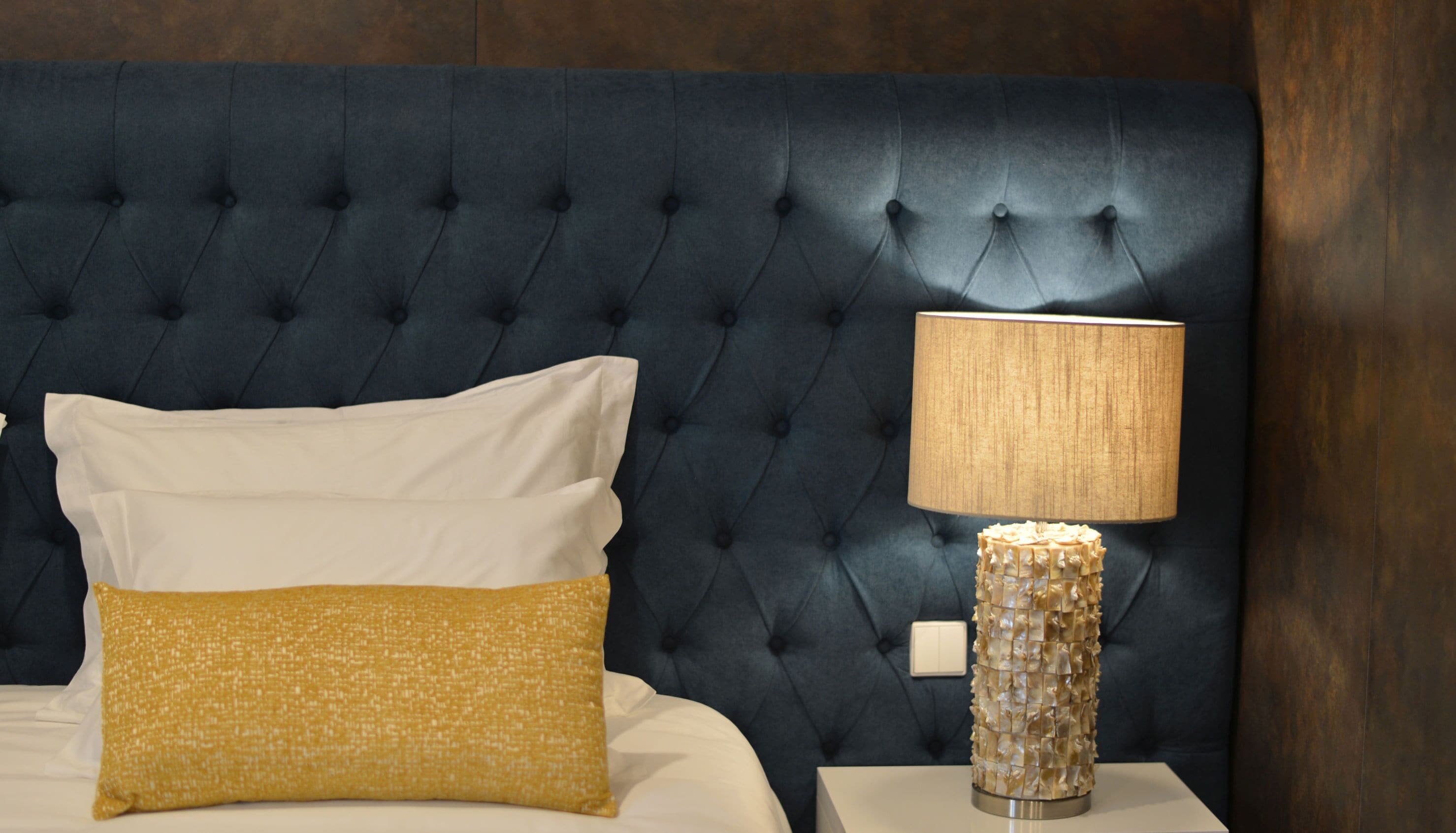 Una cama con almohadas blancas y amarillas, un cabecero capitonado azul oscuro y una lámpara de mesa con base texturizada.