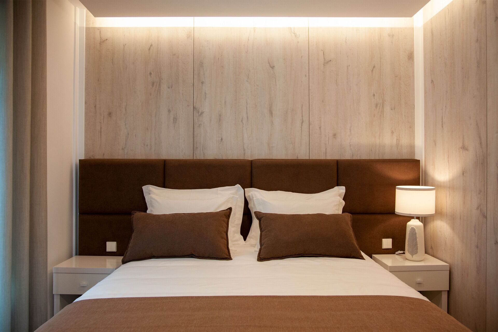 Une chambre d’hôtel avec une tête de lit en bois, un lit avec une literie marron, des oreillers blancs et une table de chevet avec une lampe et un objet décoratif.