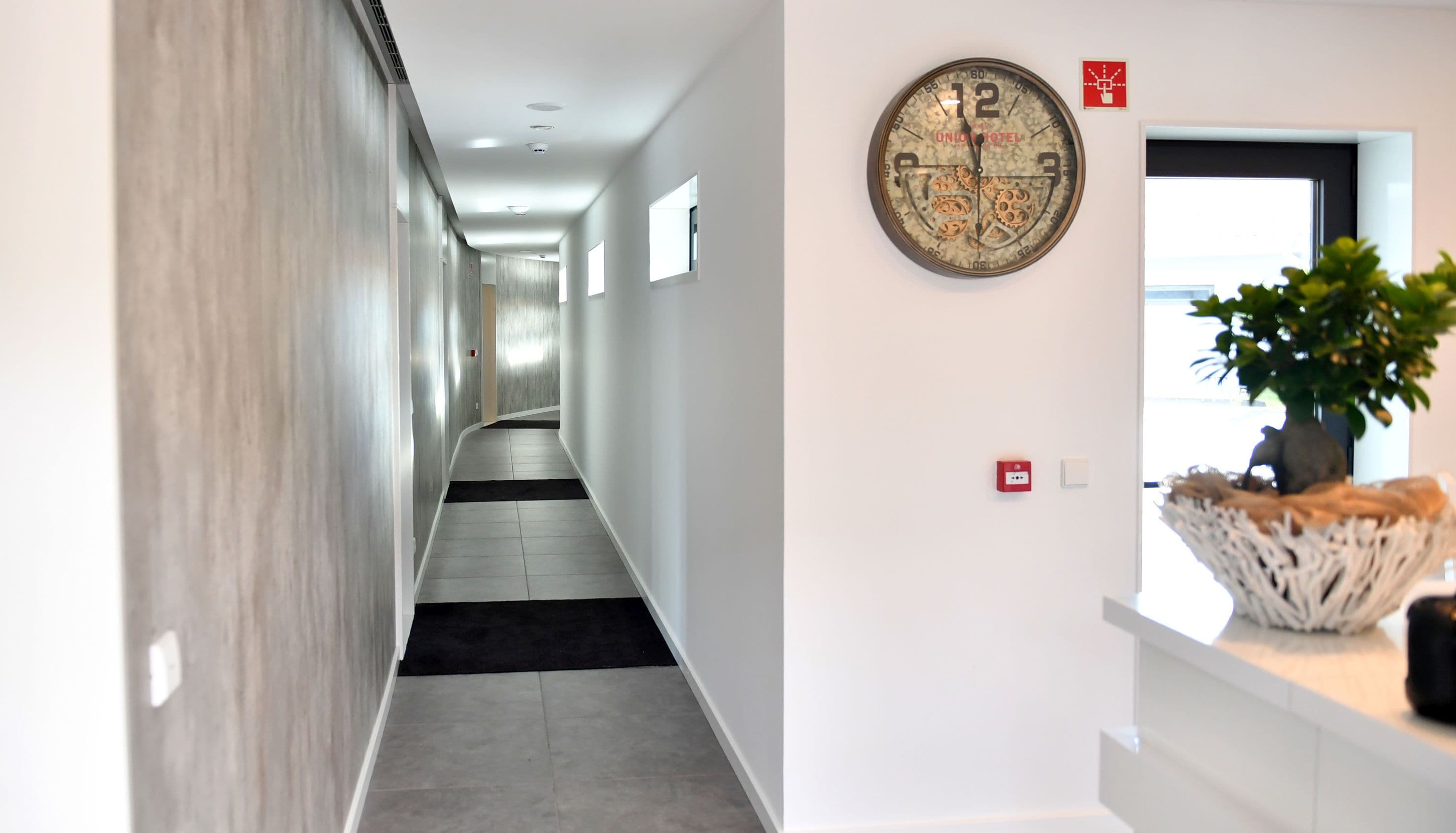 Un pasillo moderno de hotel con un reloj, una planta y un detector de incendios en la pared.