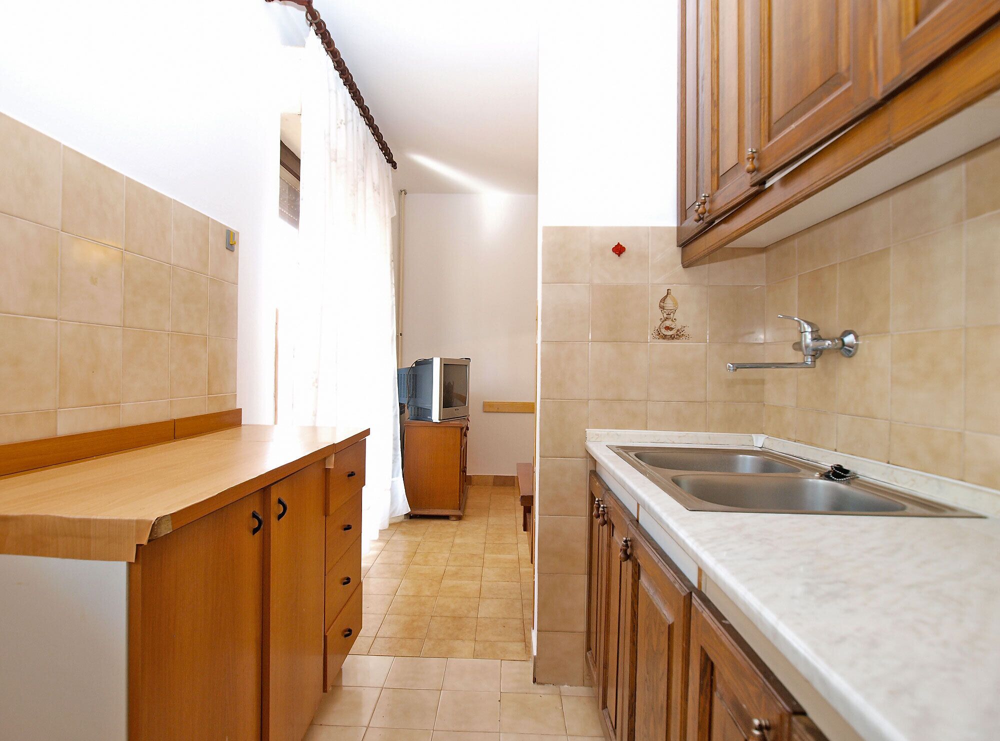 A narrow kitchen with wooden cabinets, a sink, and a small television on a stand.