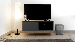A modern living room with a flat-screen TV on a wooden TV stand, a floor lamp, and a trash bin. - Midhills Premium Suites by Sparrow Homes (Genting Highlands)