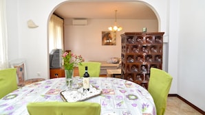 A dining area with a round table, green chairs, a floral tablecloth, a vase with flowers, and a wine bottle.