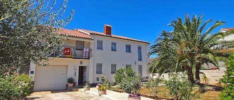 Un edificio de dos pisos con techo de tejas, un balcón con una silla roja y un patio empedrado con plantas en macetas.