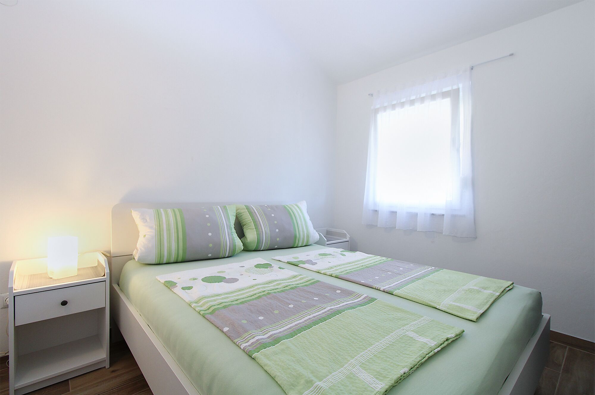 Ein Schlafzimmer mit Bett, Nachttisch, Lampe und einem Fenster mit Vorhängen.