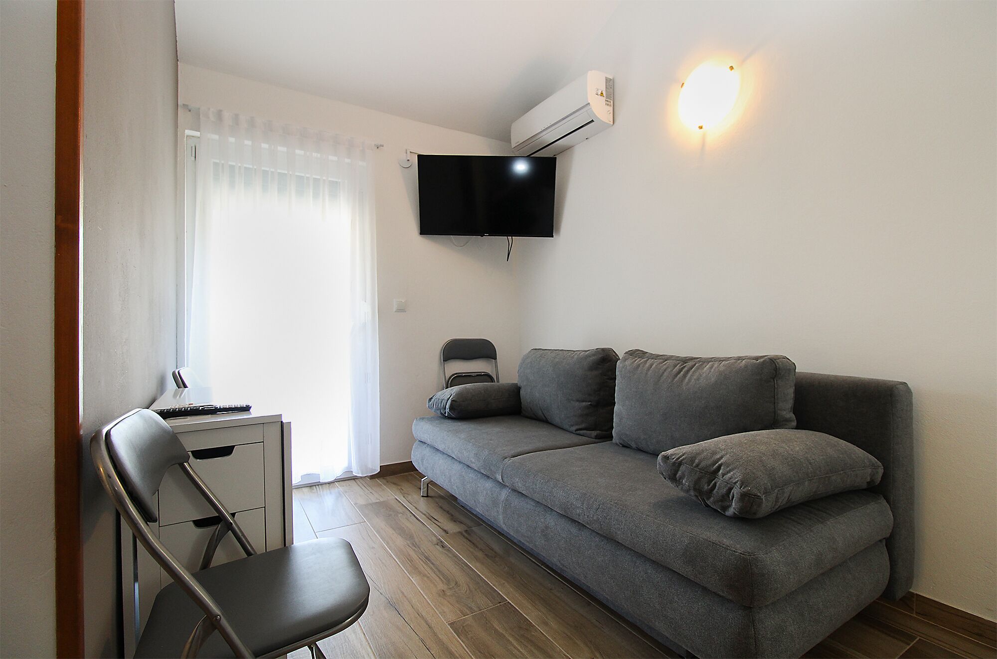 A modern living room with a grey sofa, a flat-screen TV mounted on the wall, a small desk with a chair, and a window with sheer curtains.