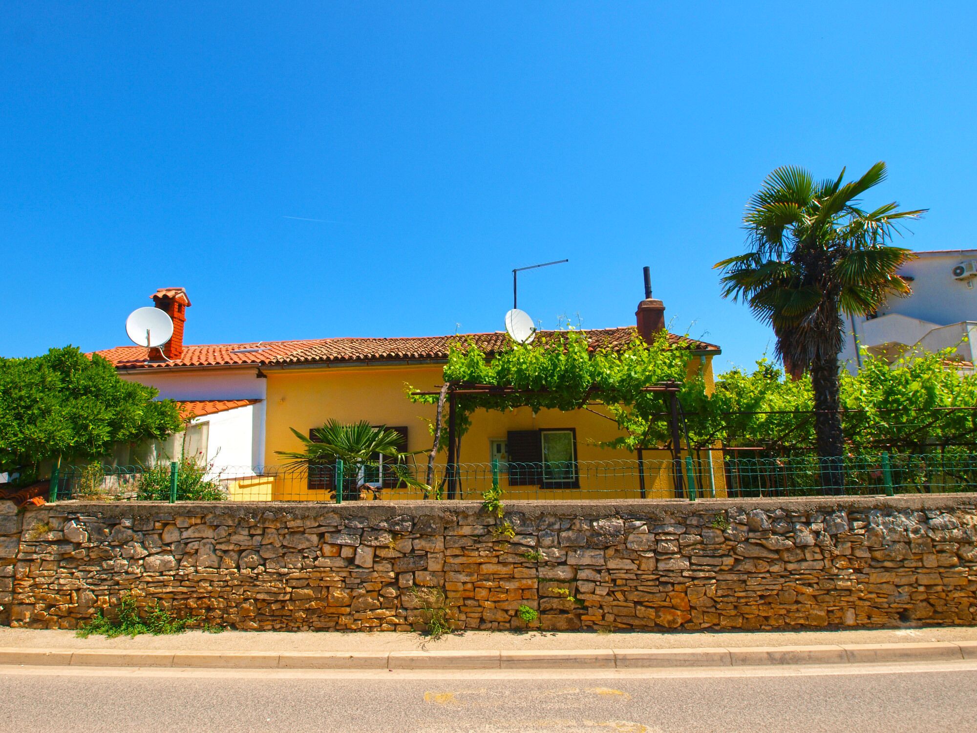 A yellow building with a stone wall in front, satellite dishes on the roof, and a palm tree to the right.