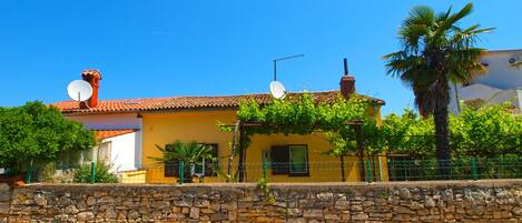 A yellow building with a stone wall in front, satellite dishes on the roof, and a palm tree to the right.