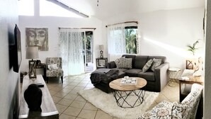 Un salon comprenant un canapé, des fauteuils, une table basse et un téléviseur. Il y a une fenêtre avec des rideaux, une porte donnant sur l’extérieur et une plante posée sur une table d’appoint.