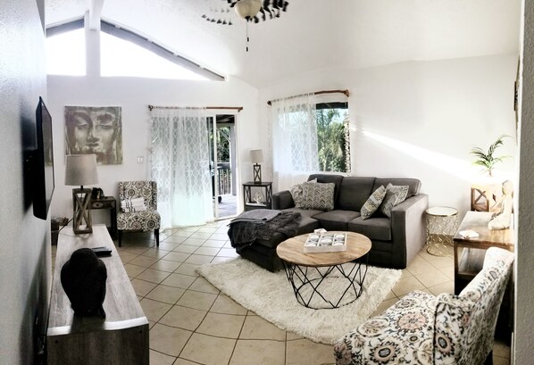 A living room with a sofa, armchairs, a coffee table, and a TV. There is a window with curtains, a door leading outside, and a plant on a side table.