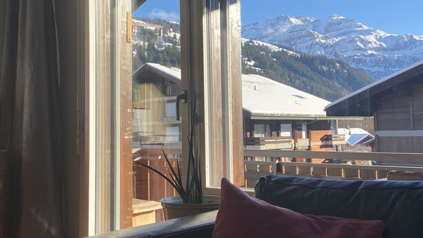 Een kamer met een groot raam dat uitzicht biedt op besneeuwde bergen en een dorp daaronder.