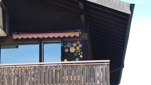 Een gebouw met een schuin dak, een balkon met houten hekwerk, en een raam met een decoratieve zon- en bloemmotief.