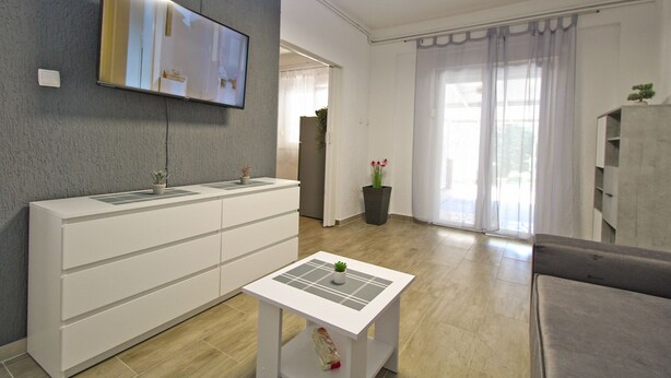 A modern living room with a flat-screen TV mounted on the wall, a white console table with potted plants, a small white table with a plant, a grey sofa, and a large window with sheer curtains. - Apartment 1052/3918 (Istria - Fažana), 300m from the beach (Fažana)