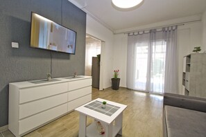 A modern living room with a flat-screen TV mounted on the wall, a white console table with potted plants, a small white table with a plant, a grey sofa, and a large window with sheer curtains.