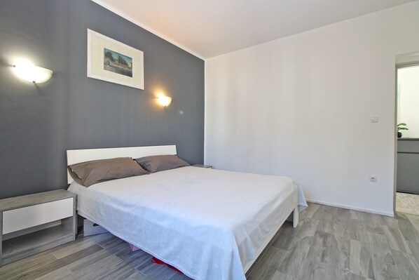 A bedroom with a bed, a bedside table, a nightstand, a wall-mounted light, and a framed picture.