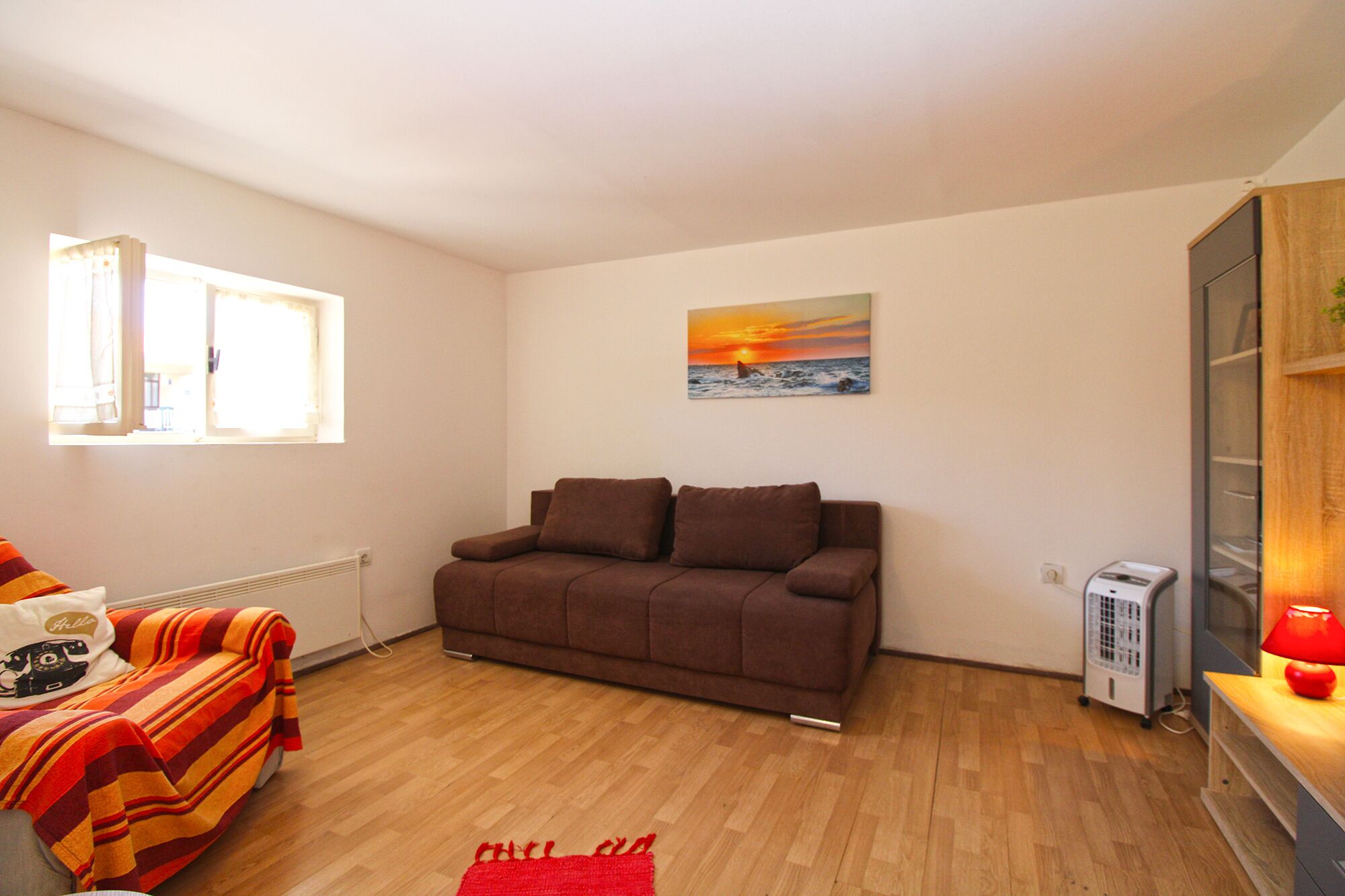 A cozy living room with a brown sofa, a red rug, a window, and a painting of a sunset.