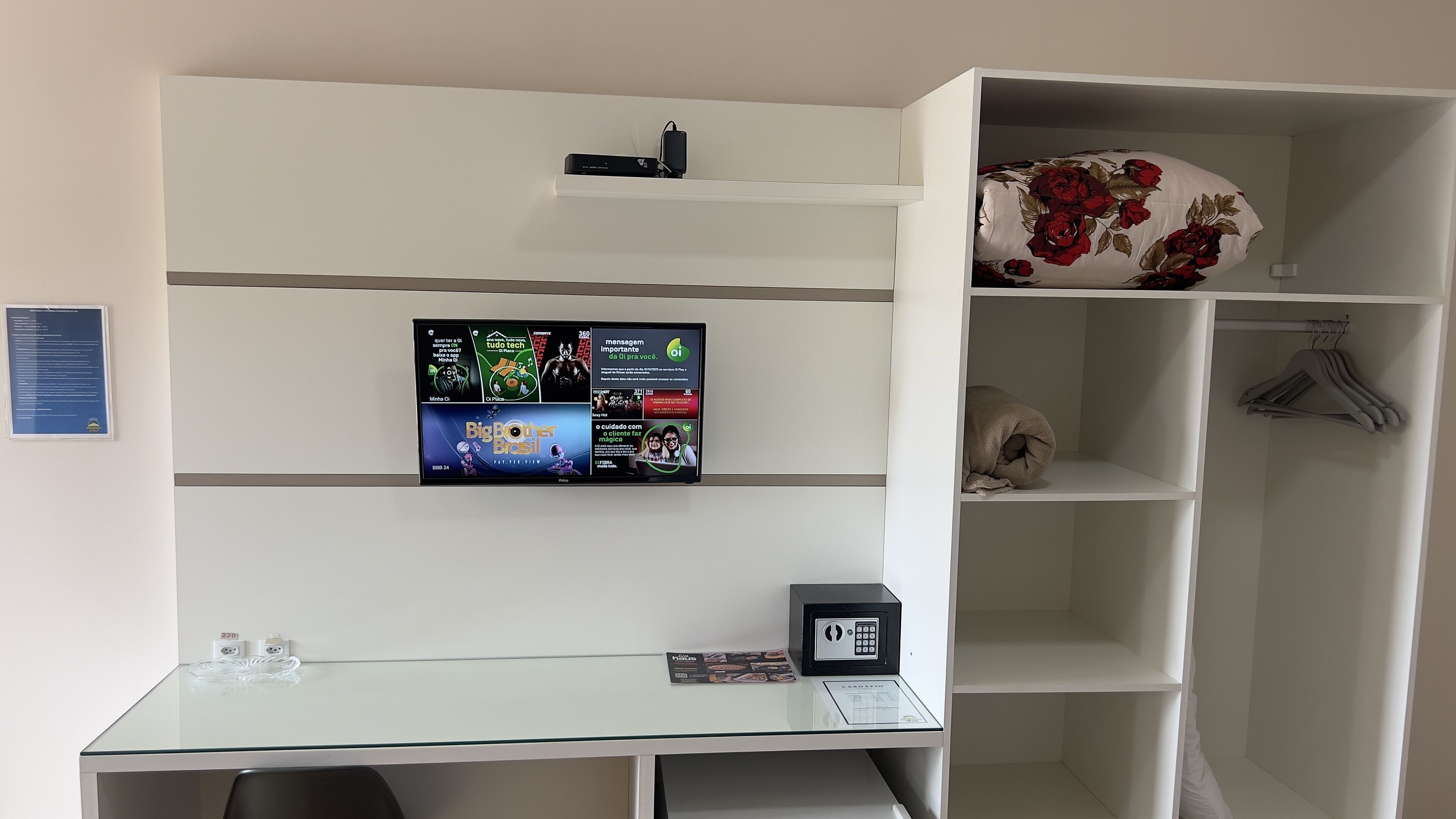 A modern room with a glass desk, a mounted TV displaying advertisements, a white wardrobe with hangers, and a neatly folded blanket.