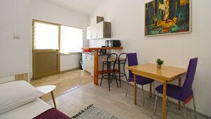 A compact kitchen with a microwave, a dining table with purple chairs, a small potted plant, and a painting on the wall.