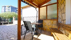 A covered outdoor seating area with a table and chairs, stone walls, and a view of buildings in the background.
