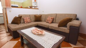 Un salon avec un canapé d’angle marron, une table basse en bois surmontée d’un chemin de table en dentelle blanche, et une petite étagère blanche agrémentée d’objets décoratifs.