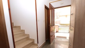 Un couloir avec un escalier en bois menant à une porte, un meuble en bois et une vue sur un espace extérieur avec un portail.