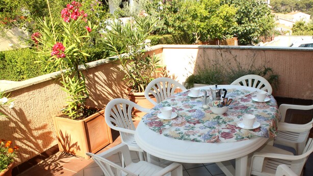 Une table ronde recouverte d’une nappe à motifs floraux, entourée de chaises en plastique blanc, est placée sur une terrasse carrelée agrémentée de plantes en pot, avec vue sur un jardin et des bâtiments en arrière-plan.