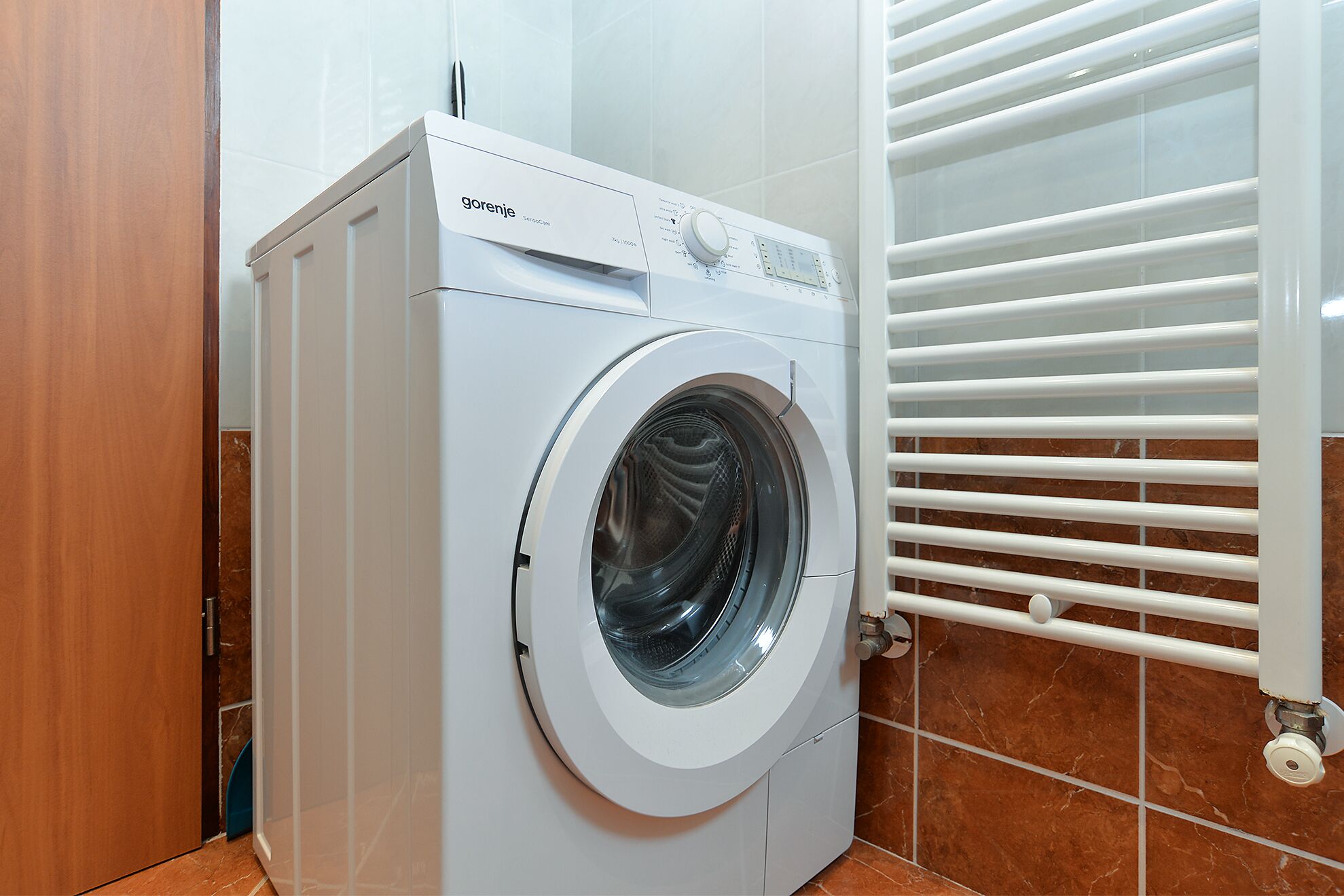 A front-loading washing machine in a bathroom with a tiled floor and a white radiator.