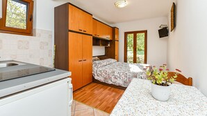 Cozinha compacta com bancada e pia, armários de madeira, cama com roupa de cama floral, uma mesa pequena com uma planta em vaso e uma janela com vista para áreas verdes.