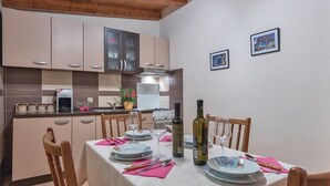 A dining area with a table set for four, including plates, glasses, and cutlery. There is a bottle of wine and a potted plant on the table. The room has wooden chairs and a kitchen in the background.