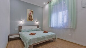 A bedroom with a bed, nightstand, wall-mounted lights, a painting, and a window with sheer curtains.