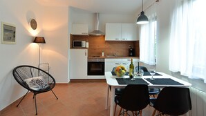 A compact kitchen with white cabinets, a microwave, and a sink. There is a dining table with chairs and a bottle of wine.