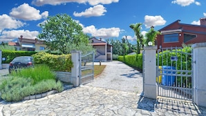 A residential area with a paved driveway leading to a gated entrance, surrounded by lush greenery and houses.