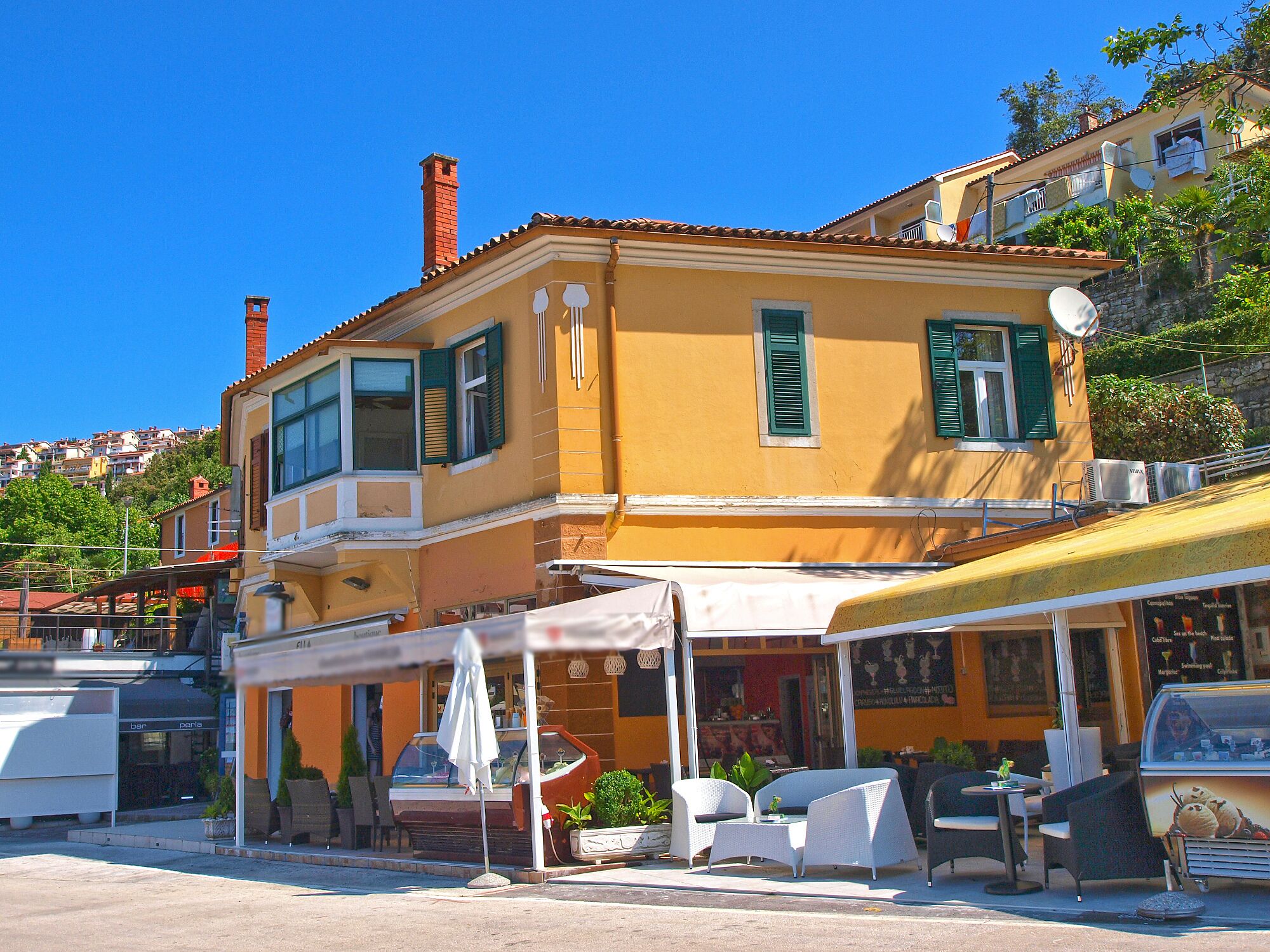 A yellow building with green shutters, a covered outdoor seating area, and a visible storefront.