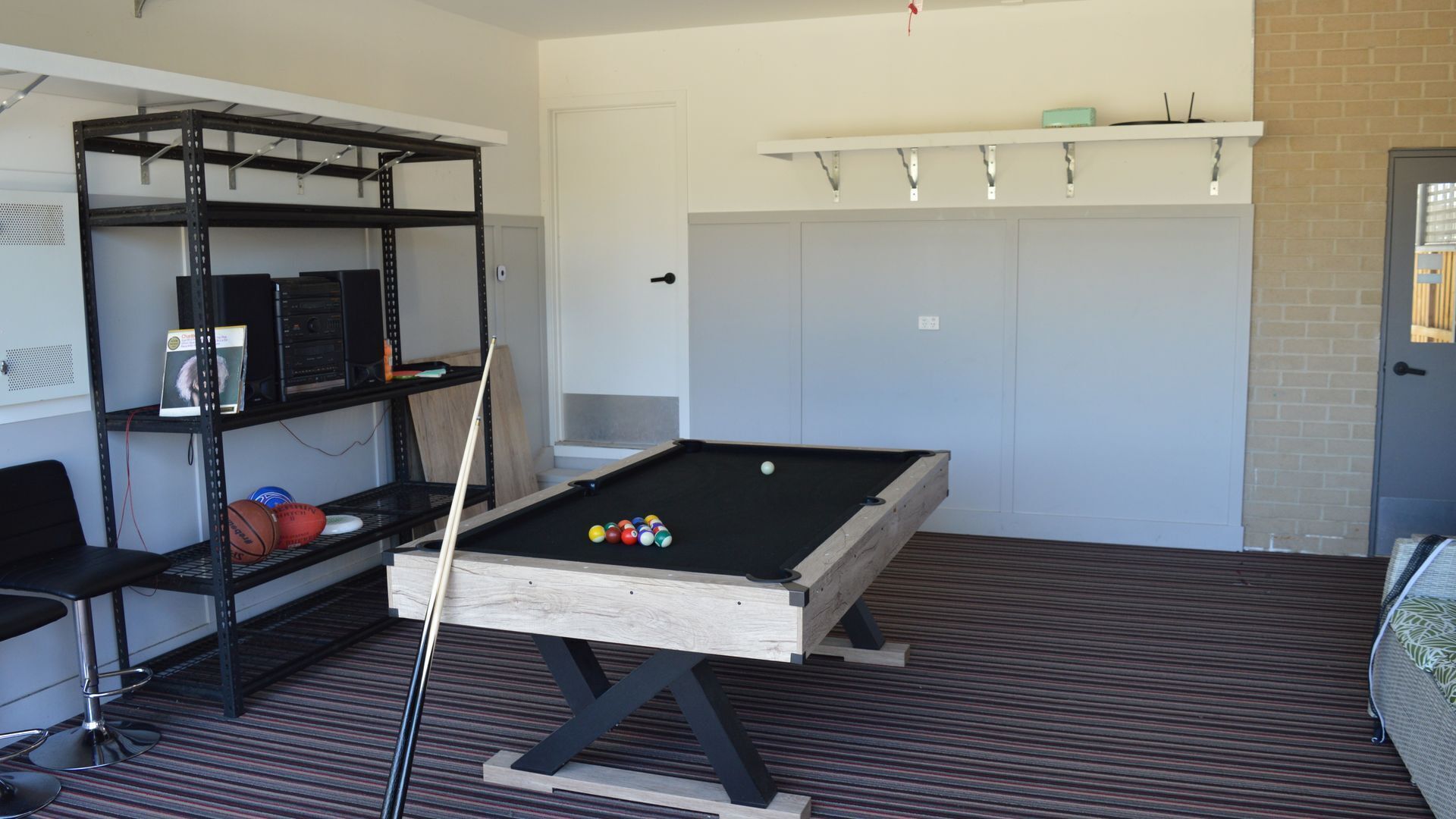 Une table de billard dans une pièce dotée d’un tapis à rayures, d’une étagère en métal et d’une porte avec une petite fenêtre.